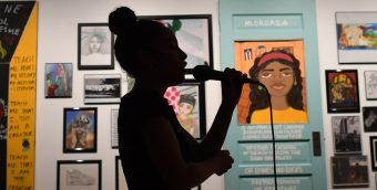 photo of a lit gallery wall, covered in works on paper and paintings. in the foreground is a black silhouette of a woman holding a mic and performing