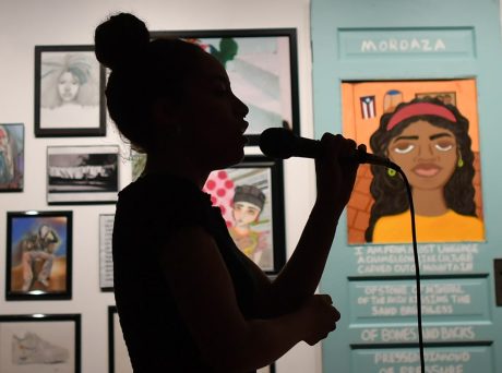 photo of a lit gallery wall, covered in works on paper and paintings. in the foreground is a black silhouette of a woman holding a mic and performing