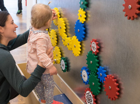 Women holds toddler up to a wall of colorful gears at the Discovery Museum in Acton.
