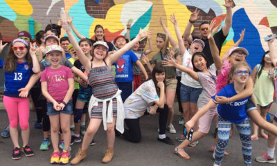 Group of young students smiling and proudly gesturing in front of a mural they helped create.