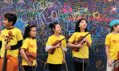 5 young people holding string instruments, smiling and standing in front of a wall of bright chalk-written phrases