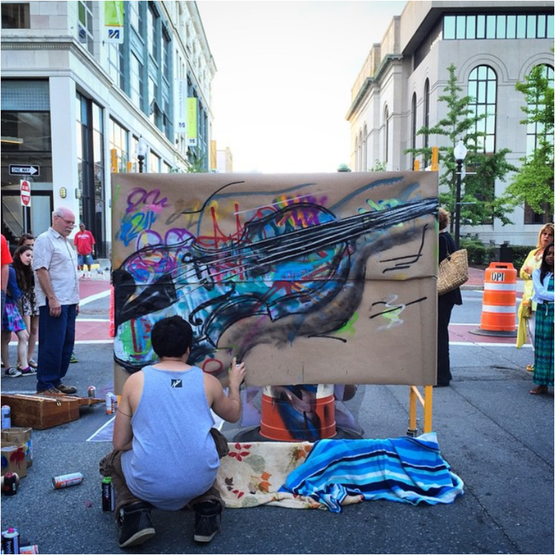 photo of a person crouching outside on a city street, holding a can of spray paint, adding a line of black to a canvas stretched across the road, outlining a painting of a colorful, street-art inspired violin
