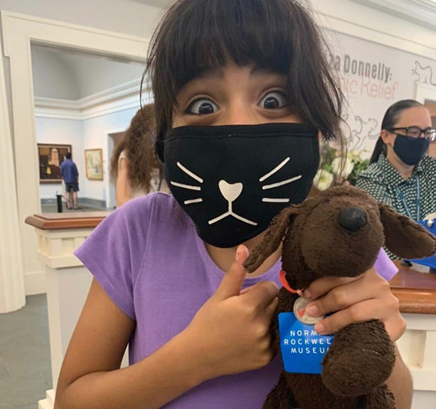 Girl wearing mask with happy eyes and a thumbs up and stuffed animal at the Norman Rockwell Museum.