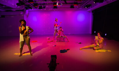 Four contemporary dancers in various poses across a stage, Pink, purple, and yellow stage lights shining on them.