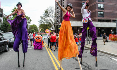three people walk on stilts, wearing bright, flowing pants, down a closed city street as part of IBA's Festival Betances