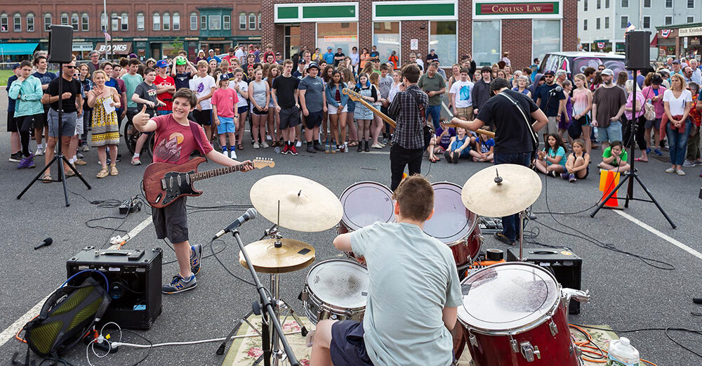 A band of four musicians play on asphalt in a parking lot as a standing crowd encircles them