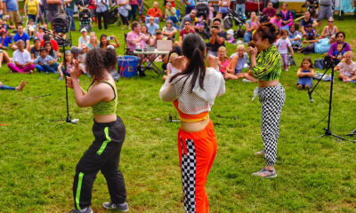 3 woman prepare to perform in front of a crowd seated in the grass in front of a row of tents