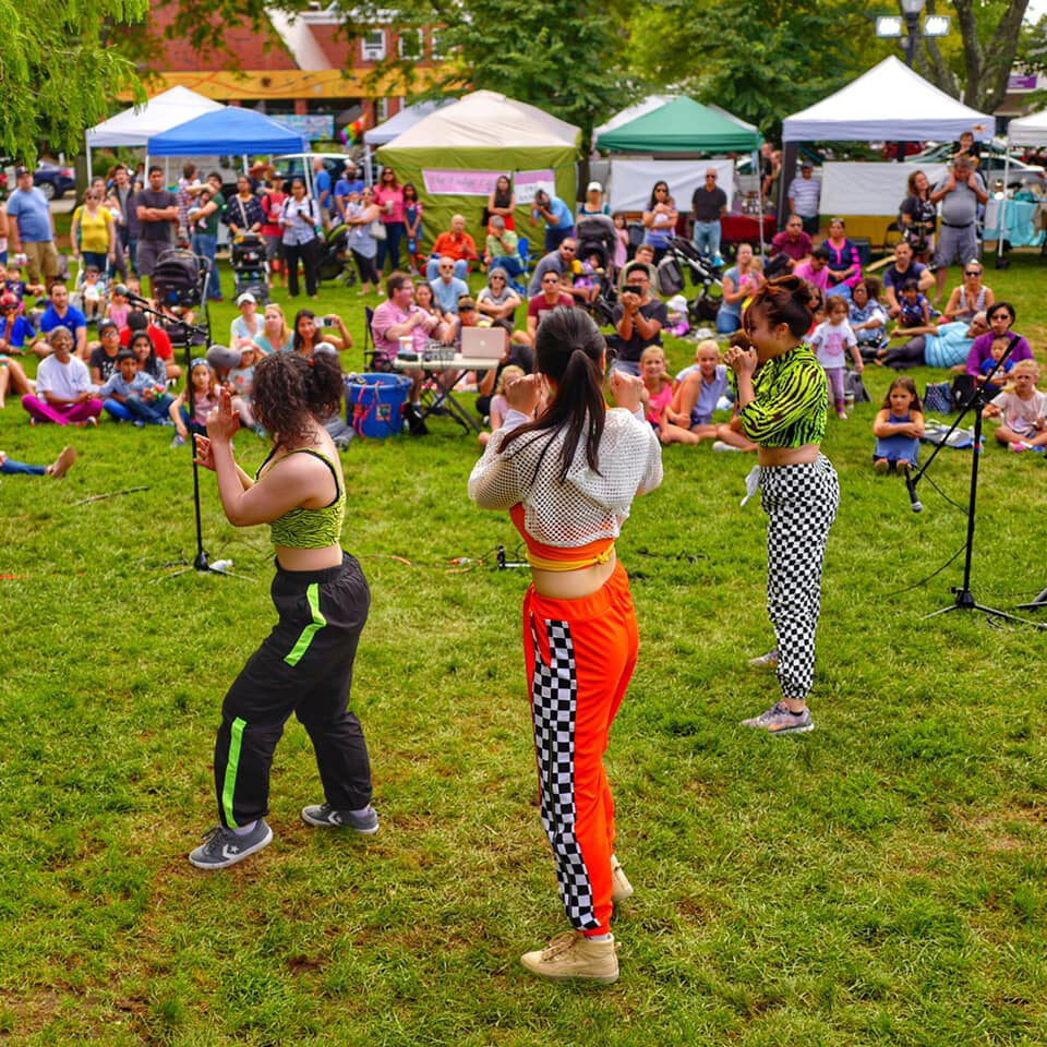 3 woman prepare to perform in front of a crowd seated in the grass in front of a row of tents