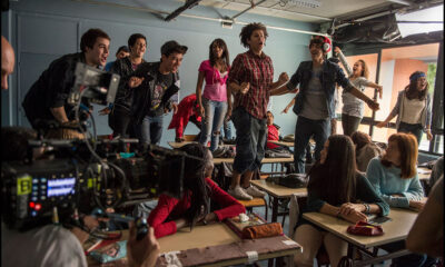 A classroom filled with students - half standing on their desks singing, the other half watching, a camera in the corner filming the action