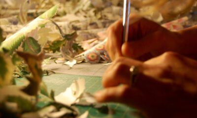 close up of an artist holding an exacto knife, cutting paper in the foreground, a pile of cut-out paper in the background