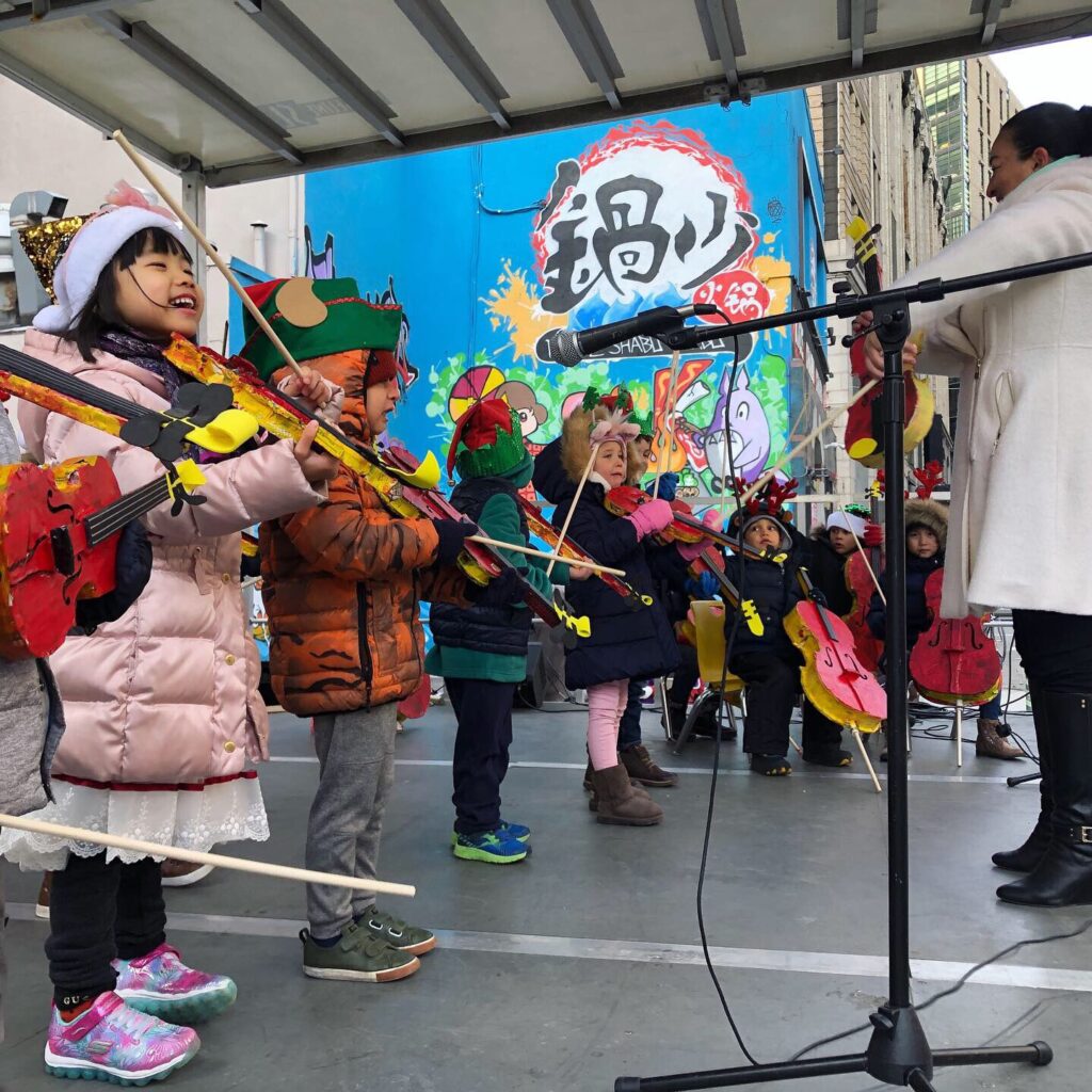 Children wearing winter hats and coats playing stringed instructments while performing outside