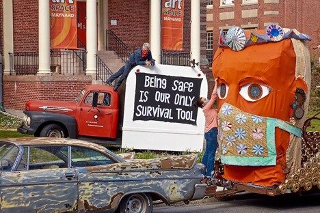 a pickup truck with a sign reading "being safe is our only surival tool" next to a 10ft tall sculpture on a trailer of a head wearing a bejeweled crown and floral cloth mask