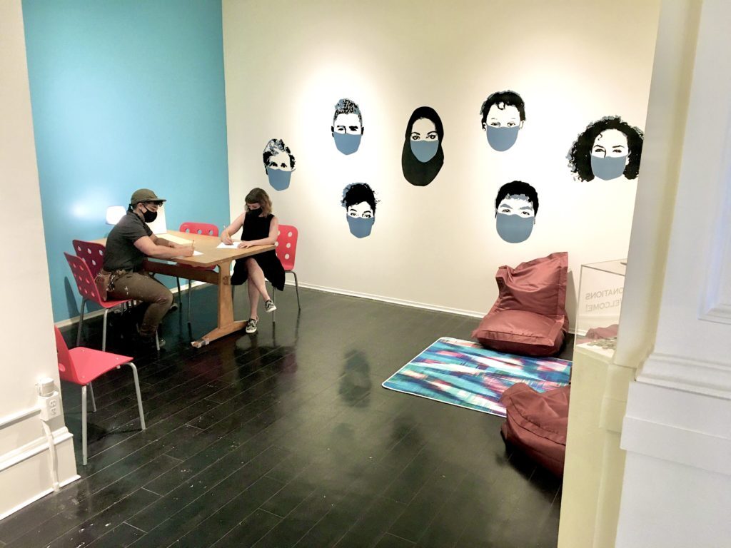 two people wearing masks sit at a table in a gallery space decorated with a mural of faces with masks