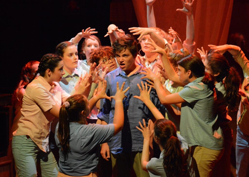A still from a theater production in which a boy is facing forward while a circle of other young actors around him have there hands open around his head