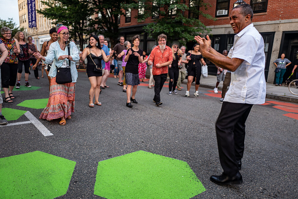 A band leader encourages a group of concert goers to dance with him in the street at a summer performance.