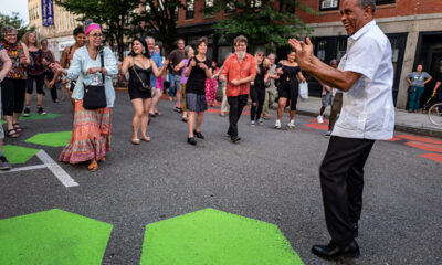 A band leader encourages a group of concert goers to dance with him in the street at a summer performance.