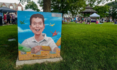 Painted utility box featuring a smiling child and grasping hands in the foreground, numerous people enjoying an outdoor event in a grassy common.
