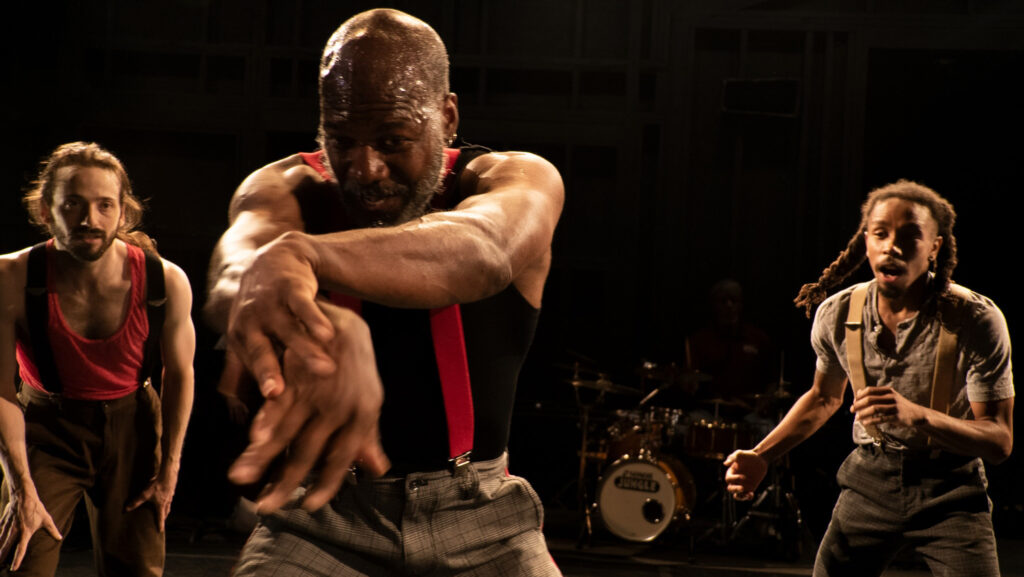 A bald male dancer with a beard wears a black sleeveless t-shirt and red suspenders. He's looking at his extended arms that cross at his hands. Two other dancers watch from the behind him.