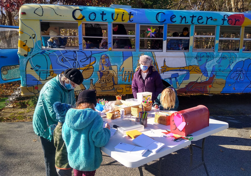 a family wrapped in winter clothing makes art on a table outside in front of a multi-colored bus