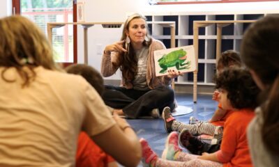 A seated woman at center of the photo surrounded by toddlers is holding up a book to display an illustration of a frog. Photo credit: Discovery Museum.