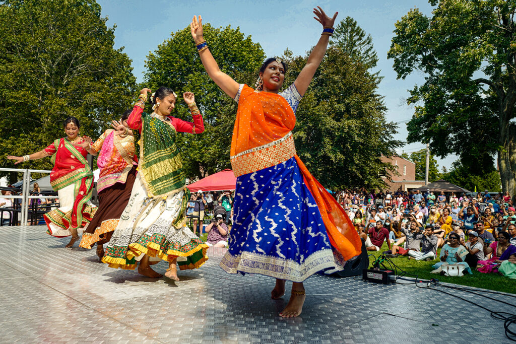 a row of dancers spin on an outdoor stage in front of a large audience seated in the grass