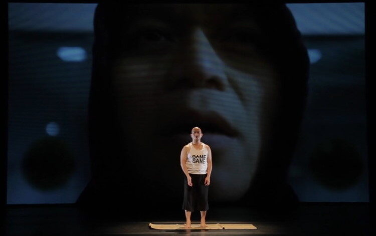 a bald dancer stands on a yoga mat on a stage behind them is a large projection of a hooded figure