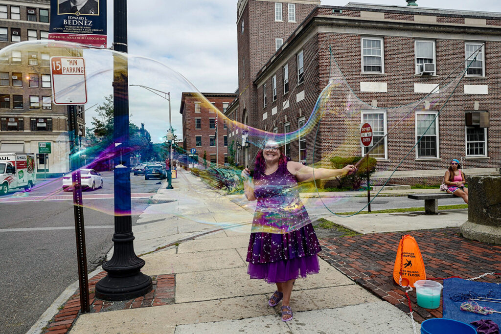 Person wearing a sparkly purple dress with arms extended outward stands on a sidewalk in Haverhill. They are holding a huge bubble wand, with a bubble wider than the sidewalk hanging in the air in front of them.