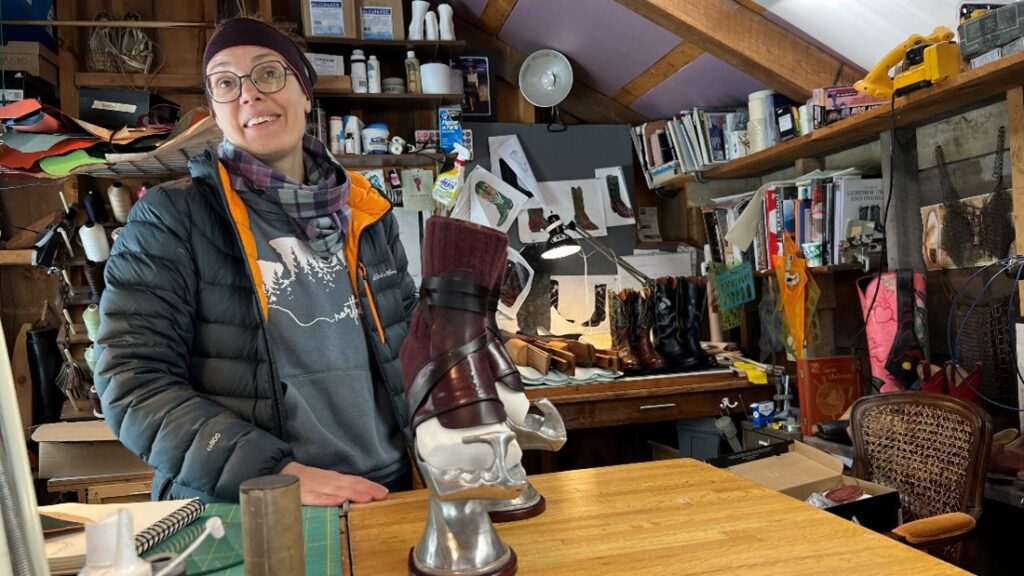 boot maker standing behind a boot mold on a worktable. All around are raw materials and sketches and other hand-crafted boots