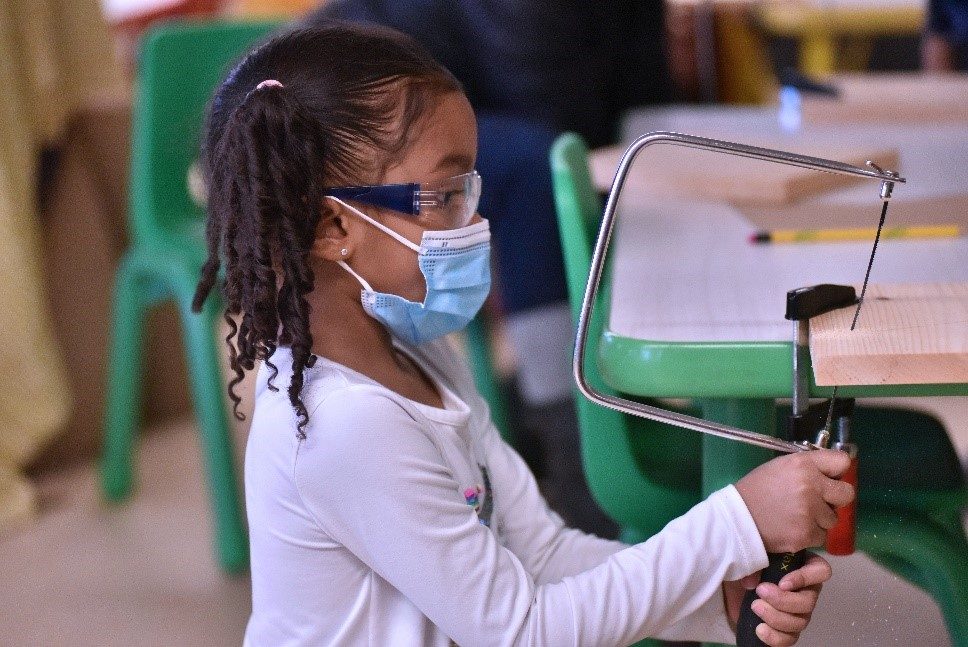 grade school student wearing safety google and a mask cuts wood using a coping saw