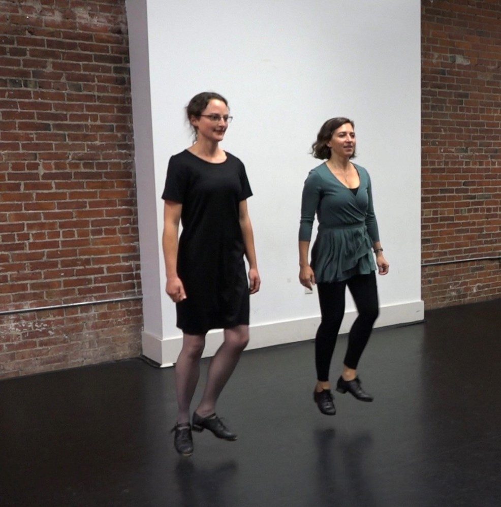 two women Irish Step Dancing indoors before a brick wall