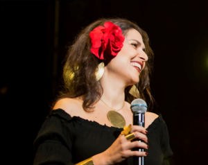photo of a brown haired woman holding a microphone and smiling during a performance. She's wearing a red flower in her hair and a black off-the-shoulders top