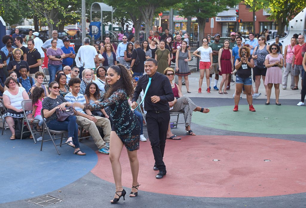 2 dancers perform on a painted playground surface surrounded by people seated and standing watching them