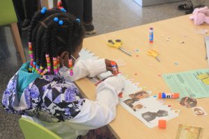 Child sitting at a table gluing images of prominent people of color to a crown-shaped piece of paper that's adorned with sparkly 'gems'