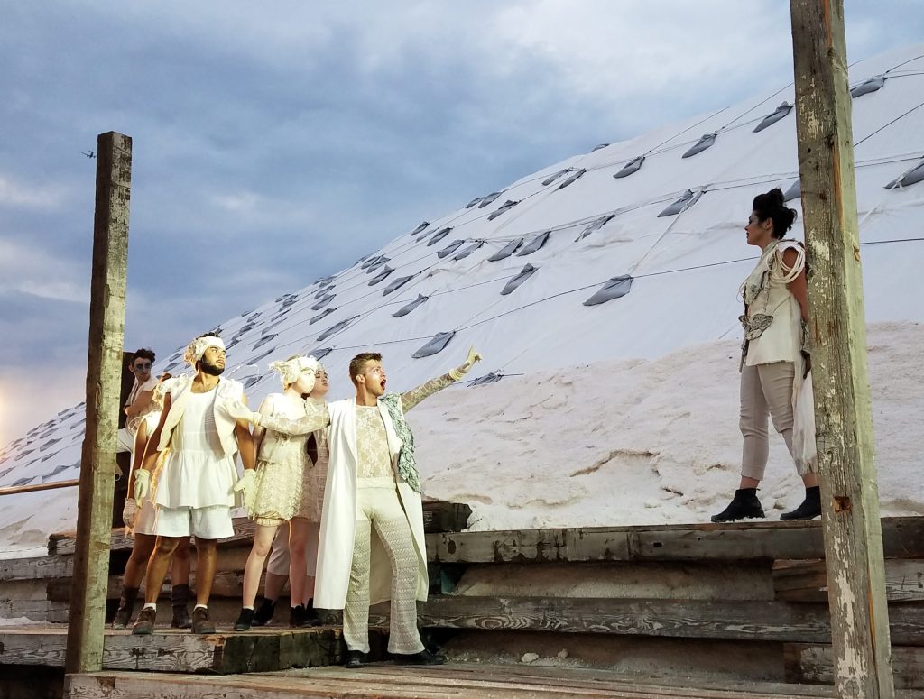 photo of an outdoor theater performance - a group of actors wearing shades of white are stopped in their tracks looking slightly up. They've come across another actor a few steps away from them and on higher ground