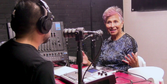 photo of a person with short, pink hair gesturing with their hands sits in a radio studio with a guest whose back is to the camera. Both people are speaking into microphones.