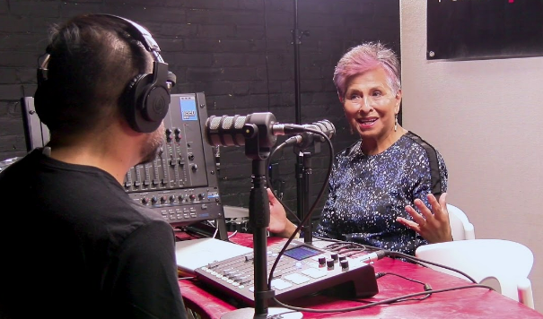 photo of a person with short, pink hair gesturing with their hands sits in a radio studio with a guest whose back is to the camera. Both people are speaking into microphones.