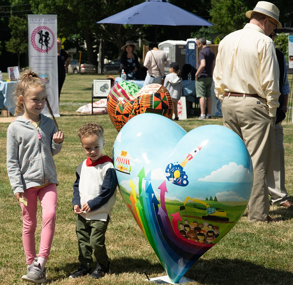photo of 2 children standing next to a heart-shaped piece of public art with a rainbow of arrows pointing up painted on it