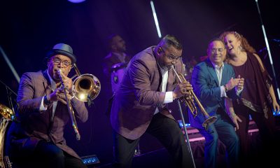 photo of a performance where a trumpet player and trombone player are playing while other band members smile and watch