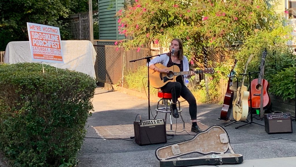 photo of a musician sitting on a stool in a driveway, singing into a microphone and playing a guitar