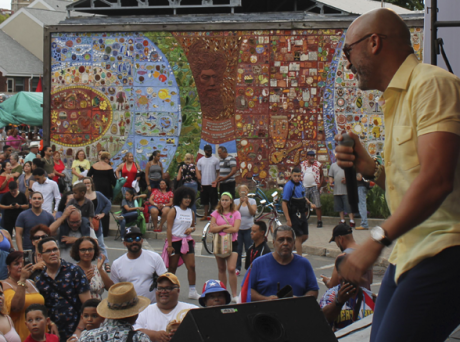 photo of performer on stage holding a mic and wearing a yellow shirt, smiling over a crowd of standing audience members. A large tile mural is in the background.
