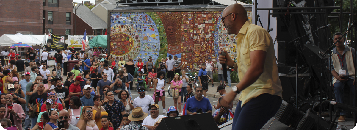 photo of performer on stage holding a mic and wearing a yellow shirt, smiling over a crowd of standing audience members. A large tile mural is in the background.
