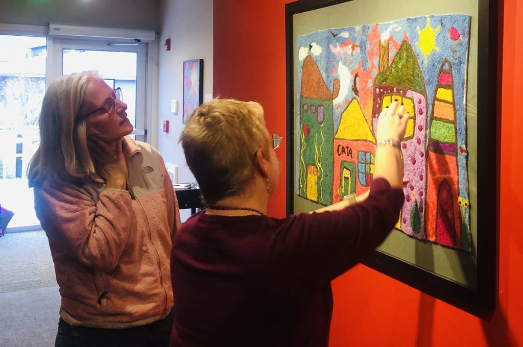 two people stand looking at a 2 dimensional work on paper consisting of colorful abstract buildings. One person has their arm raised pointing to something in the artwork.