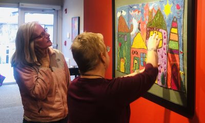 two people stand looking at a 2 dimensional work on paper consisting of colorful abstract buildings. One person has their arm raised pointing to something in the artwork.