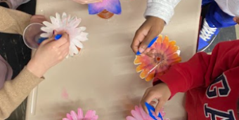 a photo taken from an aerial point of view over a school table with 5 children's sets of hands are working on a project with beads and silk flowers