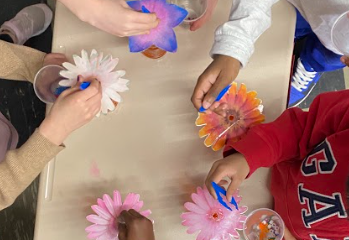 a photo taken from an aerial point of view over a school table with 5 children's sets of hands are working on a project with beads and silk flowers