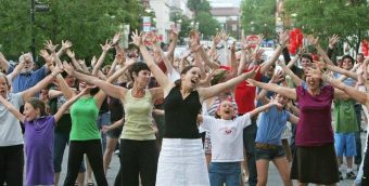 crowd of people dancing in sync outside on a roadway, their arms all uplifted