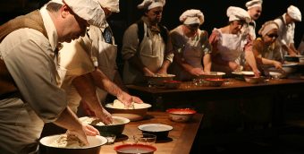 photo of a darkened stage, a long row of tables flanked on one side by 9 actors wearing white chef's hats and aprons. They are each needing dough in a bowl with an additional bowl of water in front of each person.