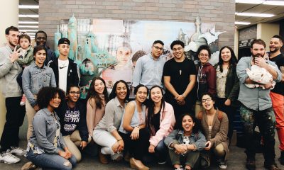 group photo of youth artists and other community members in front of a mural hanging on a brick wall in a library.
