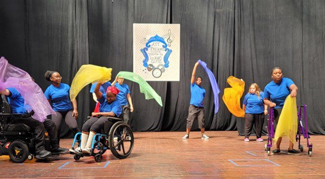 photo from a theater production 8 performers, waving larger pieces of loose fabric. Some are standing. Some are in wheelchairs. And one performer is using a walker.