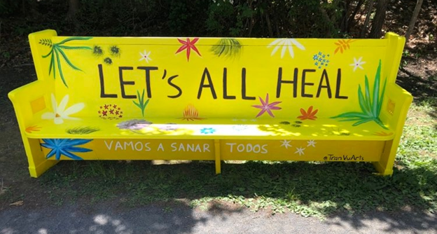 photo of a former church pew outside painted bright yellow with various flowers and leaves and the works "Let' All Heal"